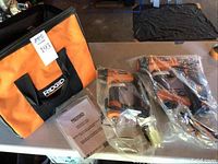Photo showing two Ridgid pneumatic nailers sealed new in plastic bags alongside a Ridgid branded orange and black carrying bag and instruction manuals on a table.
