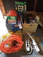 Photo showing two boxes containing multiple orange extension cords, white and black power strips, and boxed surge protector units.