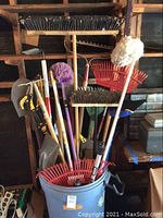 Wide view showing multiple tall garden tools including shovels, rakes, brooms, and mop arranged in and around a large grey trashcan with lid.