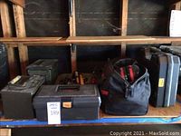 Photo showing two green metal military boxes, a metal toolbox, two tool bags and a black hard plastic DeWalt drill case on a shelf