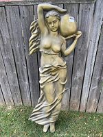 Full view of the sculpture showing the figure of a woman holding a jug, standing approximately five feet tall, in gold finish, against an outdoor wooden fence background.