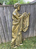 Full-front view of the gold relief wall sculpture of a woman holding fruit under a cloth, showing the size and details of the piece against a wooden fence background.