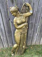 Full view of the gold relief sculpture of a woman disrobing in front of a wooden fence, showing height and overall condition.