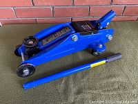 Left side view of blue trolley jack with detached pumping handle in front, showing overall condition and wheels.