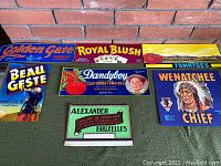 Overview of all seven lithographed crate labels arranged on a green mat against a brick wall.