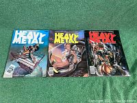 Photo showing the three Heavy Metal magazines side by side on green carpet, displaying colorful covers with sci-fi/fantasy artwork.