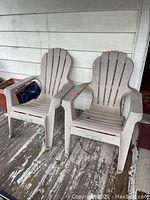 Two beige plastic Adirondack chairs placed side by side on a weathered wooden porch floor, showing slatted backrests and seats.