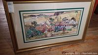 Framed art print featuring a colorful village landscape scene with trees and cottages, displayed leaning on floor, showcasing the full artwork, matting, and gold frame.