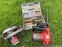 Photo shows the electric chainsaw (red and black) and the circular saw (metal body) on grass with hammers plastic case nearby