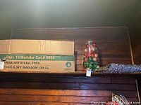 Cardboard box labeled Christmas tree Madison 6.5 ft with plastic container of assorted red and gold ornaments and coiled silver garland on wood shelf.
