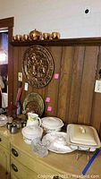 Photo of copper teapot and cups on upper wall shelf, metal wall plaques on wood-paneled wall, and assortment of ceramics and serveware on green sideboard.