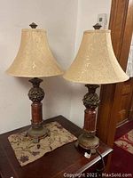 Two matching decorative table lamps placed on a wooden table with fabric mat beneath, lampshades are cream with subtle floral embossing, ornate bronze-like bases with reddish-brown cylindrical sections.