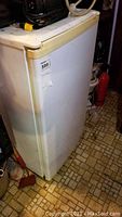 Photo showing front side of white mini fridge with yellowing on the handle area, placed on a patterned linoleum floor.