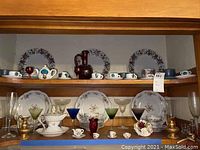 Front view of two wooden shelves containing various miniature tea sets, plates, cups, and diverse glassware including colored goblets and a dark red glass pitcher.