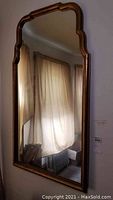 Full view of the rectangular wall mirror with a bronze tone frame showing a scalloped decorative edge hung on a wall reflecting a window and curtain.