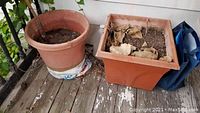 Two large reddish-brown plastic planters on a worn wooden porch floor; one round planter and one square planter containing soil and dried leaves.