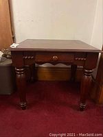 Front angle view of side table showing drawer and turned legs on a carpeted floor by a wood baseboard and white wall.