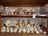 Wide view of shelves showing top shelf with China plates, clear glasses, and gold-accented teacups and saucers, and bottom shelf with miniature tea sets and large decorative ceramic pitchers and jars.