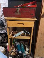 View of the red metal toolbox on a wooden cart with assorted hand tools, paint rollers, and a caulking gun, showing the collection of items in the lot.