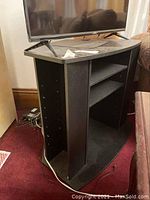 Black corner TV stand shown from an angled side view with TV on top and visible scratches on surface. Shelves are adjustable with peg holes on side panels.