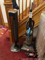 Photo shows a black and brown 39-inch tower fan and gray-blue upright Bissell vacuum with clear dirt canister in front of staircase.