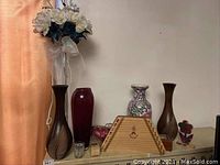 Full view of vases, music box and musical instrument arranged on shelf, features clear glass vase with faux flowers, dark wooden vases, porcelain floral vase, red glass vase, and wooden music box with Nepenorokya instrument.