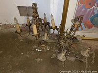 Two vintage metal chandeliers placed on a dirt floor, showing detailed view of multiple arms and candle-style bulbs in various conditions.