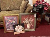 View of three floral prints leaning against couch with faux floral arrangement to the right, placed on burgundy carpet.