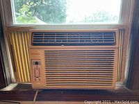 Front view of Sylvania window AC unit installed in a window, showing control panel on left and vent grilles center and right. Unit is off-white with yellow side panels.