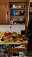 Kitchen countertop with multiple wooden bowls and a few larger serving bowls between a red enameled pot and a smaller brown pot. Shelves above containing miscellaneous kitchen cups and mugs.