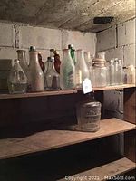 Wide view of shelf with collection of old glass bottles and milk jugs of various sizes and styles, some with caps, some empty, dusty with age.