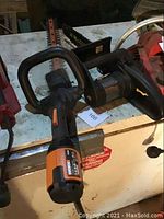 Shows the electric hedge trimmer with black and orange handle and long trimming blades.