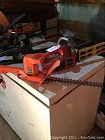 Two Black and Decker garden tools placed on a white cabinet: an orange electric hedge trimmer in front and a black gas powered chainsaw behind it.