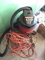 Craftsman red and black shop vacuum with flexible hose and orange extension cord tangled around it.