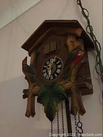 Front view of wooden cuckoo clock showing clock face with Roman numerals, two carved birds on sides, and leaf decoration below clock face.