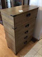 View of the 5 drawer wooden dresser showing top and front with metal corner brackets and handles, floor visible beneath.