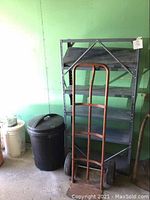 Photo showing metal shelving unit with five shelves, a hand dolly leaning against it, and a black garbage bin nearby.