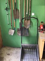 Photo showing a wall with hanging garden tools including shovels, hoes, pitchfork, and a large black snow shovel on the floor in front.