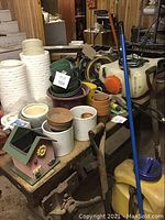Full lot showing stacked white and green plant pots, clay pots, yellow fertilizer sprayer, birdhouse, shovels and broom on and under the table.