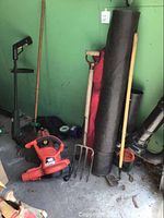 View of the gardening tools and equipment clustered near a green wall including blower vacuum, pitchfork rake, hoe, rolled fabric, and gas can.
