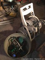 Photo shows a gray plastic portable hose reel with garden hose coiled around it, and a metal bucket containing sprinklers and hose connectors.
