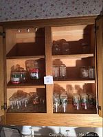Wooden cabinet with multiple shelves holding various sets of glasses including small clear glasses and decorated glasses.
