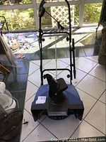 Full view of black electric snow thrower with upright handlebar, control cables, and wide auger housing on a tiled floor.