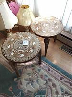 Two matching wooden round tables with carved elephant face legs and elephant inlay designs on tops, placed on a floral rug near a window with curtains. Lamps are visible nearby.