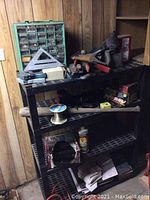 Photo showing black plastic shelving unit loaded with nuts and bolts organizer, hand tools including hammer, saw, carpenter's square, spool of wire, and various hardware items.