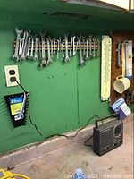 Photo showing a rack with multiple metal wrenches hanging and a black portable AM/FM radio on the floor of a workshop area with green walls.