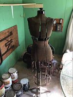 Full view of vintage dress form mannequin standing on three-legged metal base in a workshop-like room, showing the fabric torso and metal cage lower half.