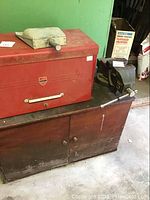 Wooden cupboard with two doors and round wooden handles, wheelchair visible in background; red metal toolbox on top of cupboard surface; black metal vise mounted on right side of cupboard.