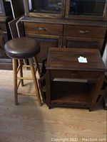 Wooden end table with drawer and open shelf next to wooden stool with padded leather seat, from side angle