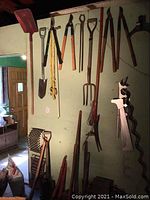 Photo showing a variety of hanging yard tools including shovels, branch cutters, axes, pitchfork, and long saw blades along green wall in workshop.